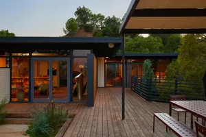 A modern house with a wooden deck and patio surrounded by trees, featuring a large window, TV, and natural sunlight