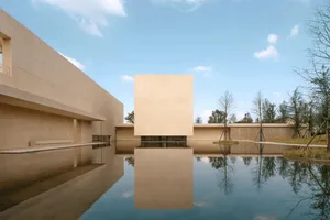 A modern building with a large glass wall and a reflecting pool in front, surrounded by natural sunlight and a calm atmosphere