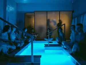 A group of people sitting on steps and chairs around a large pool with a blue tint, enjoying a relaxed and social atmosphere