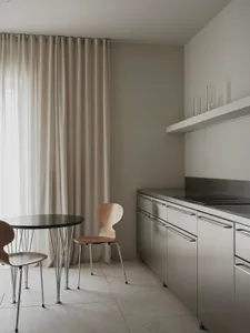 A modern kitchen with a dining table and chairs, surrounded by white surfaces, with natural light and a sink in the background