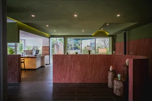 A hotel lobby-style room with a red accent wall, a green ceiling, and a counter with a window behind it, featuring two potted plants and a doorway to another room