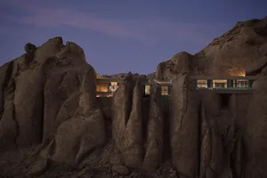 A rustic stone house built into a rocky mountain, set against a dramatic mountain backdrop with dim lighting