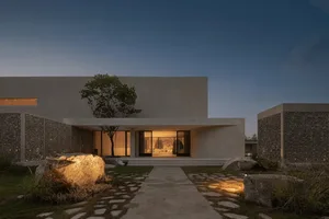 Modern building with a concrete exterior, large window, and door, set against a night sky with a tree in front, illuminated by the building's lights