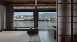 A modern living room with a bed, couch, and sink, featuring a large window with a city view, wood, and glass elements, with natural light and a bright atmosphere