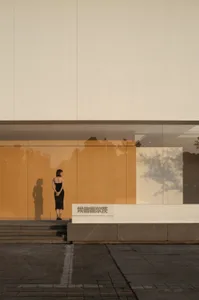 A woman stands in front of a modern building with a glass wall, her reflection visible on the glass, creating a sense of depth and elegance