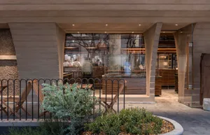 A restaurant dining area with wooden tables and chairs, potted plants, and natural lighting, creating a warm and inviting atmosphere