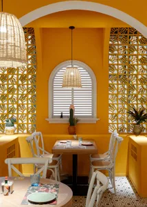 Dining area with a table, chairs, and a potted plant, surrounded by yellow walls and a window, with cups and bottles on the table, creating a warm and inviting atmosphere