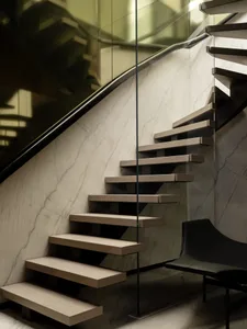 A luxurious staircase with a marble staircase and glass railing in a living room with a black chair and couch