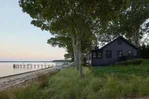 A black house with a large chimney situated near a body of water, surrounded by a grassy area and a few boats
