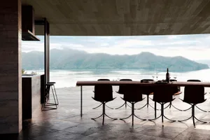 A modern dining room with a long wooden table set with wine glasses, plates, and cutlery, surrounded by chairs in a large space with natural light and a view of the mountains through the windows