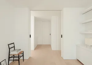 A bright and welcoming hallway with a white color scheme, wood accents, and a chair in the corner, set against a modern and clean backdrop