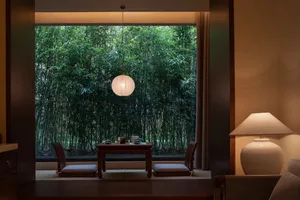 A dining room with a wooden table and chairs, a lamp, and a window with a forest view, creating a warm and inviting atmosphere