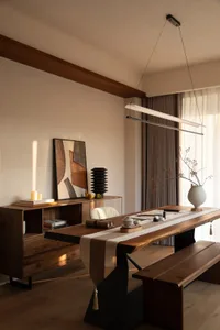 A modern living room with a wooden table and bench, set with a vase, bowl, and cups, surrounded by a couch and a potted plant, with natural light from a window creating a calm and inviting atmosphere