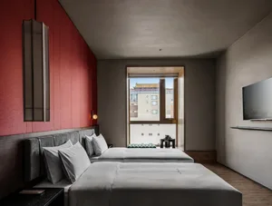 A well-lit bedroom with a bed, white bedding, and a red wall, natural light enters through the window