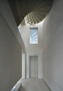 A modern hallway with a concrete spiral staircase, illuminated by natural light from a window, creating a calm and inviting atmosphere