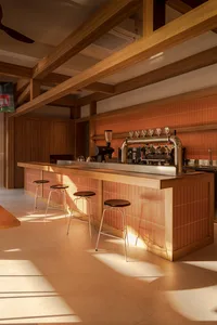 A well-lit bar area with a wooden counter, black stools, and a coffee machine in a restaurant setting, featuring natural lighting and high-quality materials