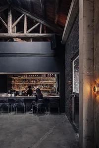 A casual bar scene with a black counter, bottles, and a woman sitting at the counter with chairs and a dining table in the background
