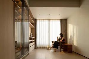 A woman sits in a well-lit living room or dining area with a wooden floor, a dining table, and a couch, surrounded by warm and inviting atmosphere