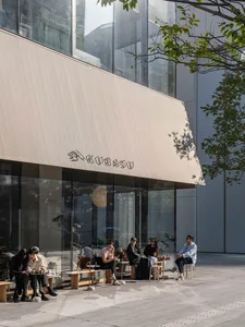 A sunny outdoor cafe scene with a white-roofed building, large window, and people sitting on chairs and benches
