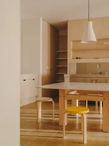 Modern kitchen with wooden floors and cabinets, featuring a sink, refrigerator, dining table, and chairs, with natural light and a warm atmosphere