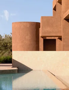A desert villa with a red brick facade, a large circular window, and a swimming pool surrounded by a brick wall and a bench