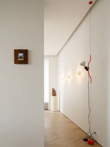 A dimly lit hallway with a white wall, a red cord, two framed pictures, a lamp, and a clock, in a minimalist style