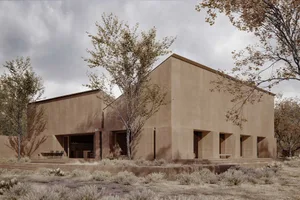 A stone building with a large window and door stands in a desert landscape with a single tree in front