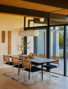 A modern dining room with a wooden table and chairs, surrounded by a large window and filled with natural light, featuring a bowl of fruit on the table