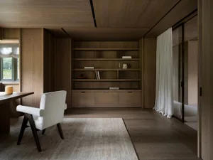 Cozy living room with wooden floors and furniture, a large bookshelf, chair, and dining table, well-lit with natural light from windows