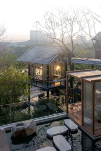 A modern house with a glass-walled living room and patio, surrounded by a wooded area, featuring a couch, dining table, pool, and natural lighting