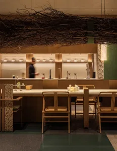 A modern restaurant dining area with a long wooden table, chairs, and vases, set with wine glasses, cups, and bowls, under warm natural light