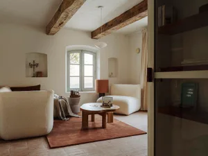 Comfortable living space with a wooden floor, white walls, a couch, chair, and dining table, well-lit by natural light through a window