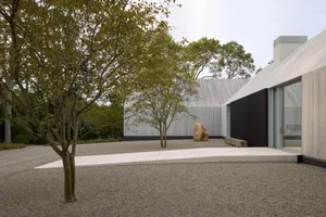 A modern house with a stone exterior and a large rock in the front yard, set in a calm and serene outdoor setting with a patio area visible