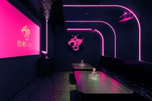 A stylish dining space with a bar, black furniture, and a pink and purple color scheme, set against a black and white backdrop, with a mix of natural and artificial lighting