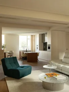 A modern living room with a white couch, chair, and dining table, featuring a kitchen area with a refrigerator and oven, and natural light from windows