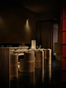 Dimly lit dining room with a wooden floor, a dining table, chairs, wine glasses, and a vase, creating a cozy atmosphere