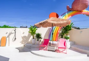 A colorful outdoor patio with a pink umbrella, chair, and bench, surrounded by a rainbow mural and lush surroundings