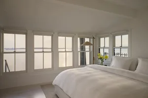 A serene bedroom with a white comforter, yellow flowers, and a wooden floor, lit by natural sunlight through the window