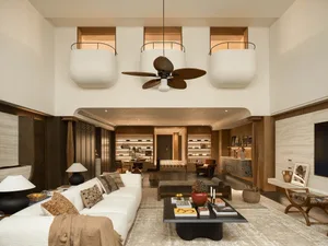 A well-lit living room with a white couch, a chair, and a dining table, featuring a large ceiling fan and a chandelier, with natural light coming in through the windows, creating a warm and cozy atmosphere
