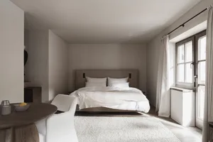 A dimly lit bedroom with a bed and chair, both white, near a window allowing natural light