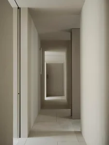 Narrow hallway with concrete walls, white color, and dim lighting, leading to an open doorway