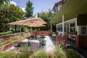 A patio with a dining table and chairs surrounded by a mix of wood and metal furniture, shaded by a large umbrella, in a natural outdoor setting with sunlight
