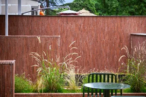 A casual outdoor seating area with a brown brick wall, a wooden bench under an umbrella, and a nearby dining table in a soft, diffused light