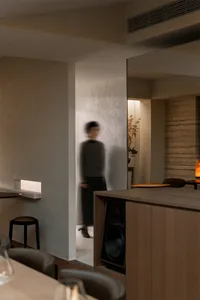 A woman walks through a modern kitchen with a wooden floor and a white wall, featuring a dining table and chair