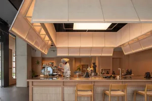 Dimly lit kitchen with a wooden counter, chairs, and dining table, featuring a coffee maker and a cup on the counter