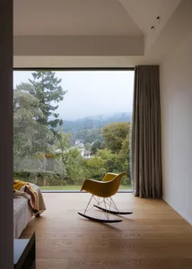 A modern living room with a bed, chair, and couch in front of a large window, surrounded by a warm and inviting atmosphere with natural light and wood materials