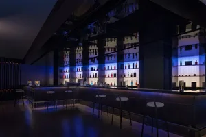 A modern bar with a blue lighting effect, featuring numerous bottles on shelves and a counter with seating for patrons