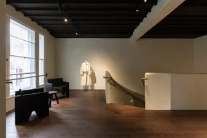 A modern lobby with a wooden floor, white walls, a staircase, and seating areas with chairs and a bench, lit by dim lighting