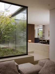 A well-lit living room with a glass wall, a couch, a chair, and a dining table, featuring a modern and minimalist design