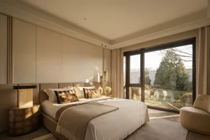 A well-lit bedroom with a bed near the window, a chair nearby, and a beige color scheme creating a warm atmosphere